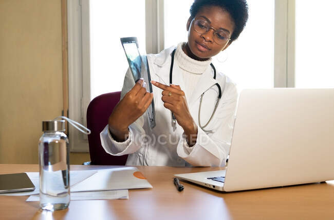 Médico femenino étnico que señala la imagen de rayos X de la columna vertebral contra netbook en la mesa en la clínica - foto de stock