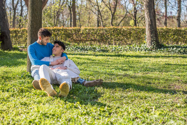 Вид збоку усміхнені пару гомосексуальних чоловіків сидять на галявині в парку і насолоджуються сонячним днем, дивлячись один на одного — стокове фото