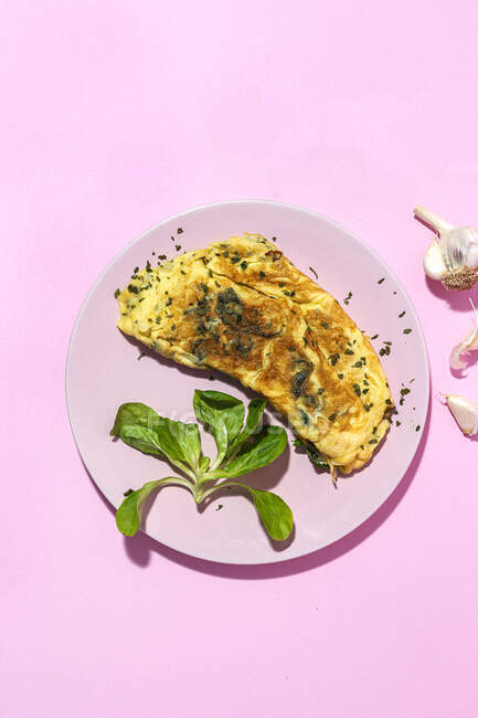 Sabrosa tortilla en plato contra ramitas de perejil fresco con dientes de ajo sobre fondo rosa - foto de stock