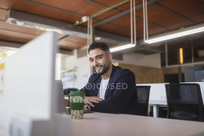 Alegre empresario masculino mirando a la cámara mientras trabaja en el lugar de trabajo sentado en la mesa con el ordenador portátil - foto de stock