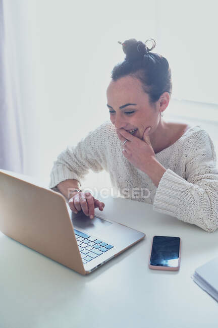 Весела жінка віддаленого працівника, що серфінгує Інтернетом на нетбуці за столом з смартфоном і копіювальною книгою вдома на сонячному світлі — стокове фото