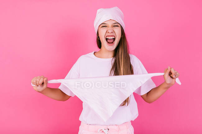 Беззаботный подросток в повседневной одежде с каштановыми волосами и платком, представляющий концепцию осознания, глядя на камеру, стоящую на розовом фоне — стоковое фото