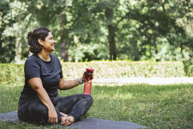 Side view of tired young lady with dark hair in sportswear sitting on mat on grassy lawn with crossed legs and drinking water after yoga training in park — Stock Photo