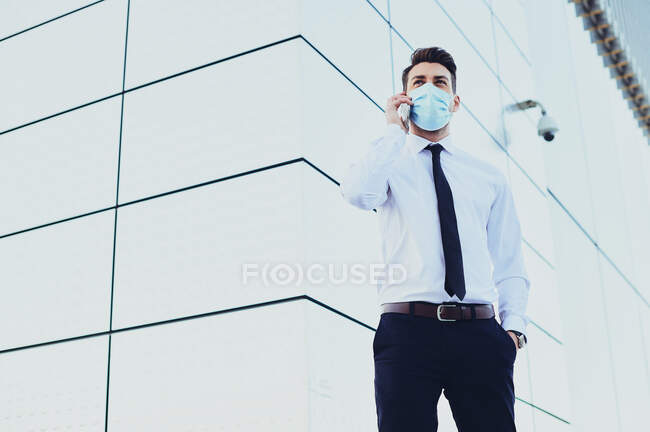 Стильний керівник чоловічої статі в офіційному одязі та медичній масці, що розмовляє на мобільному телефоні, дивлячись далеко в місто — стокове фото