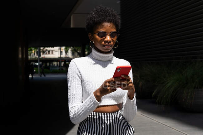 Mulher afro-americana em roupas da moda navegando nas mídias sociais no celular enquanto caminhava na rua da cidade no dia ensolarado — Fotografia de Stock