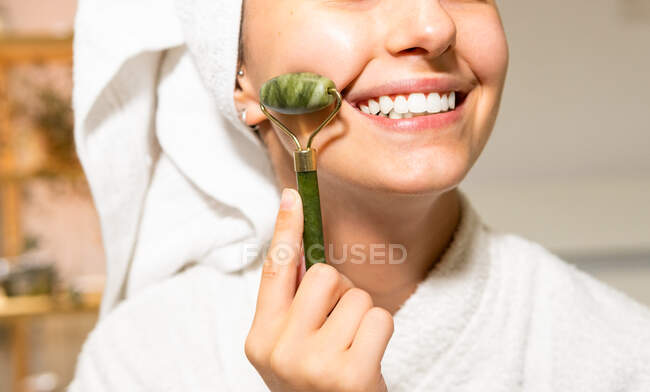 Cortar hembra joven con toalla en la cabeza sonriendo y masajeando la cara con rodillo de jade durante la rutina de cuidado de la piel en casa - foto de stock