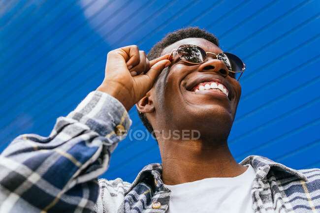 Niedriger Winkel eines gutaussehenden, lächelnden afroamerikanischen Mannes mit trendiger runder Sonnenbrille, der im Sommer auf der Straße vor hellblauem Hintergrund wegschaut — Stockfoto
