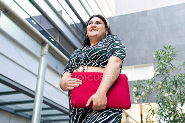 Side view of cheerful young female in stylish striped outfit with red laptop case in hands smiling while standing against contemporary city building — Stock Photo