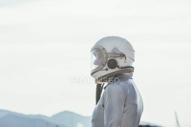 Vista lateral del hombre en traje espacial y casco mirando hacia otro lado mientras está parado en el camino en el día soleado en la naturaleza - foto de stock