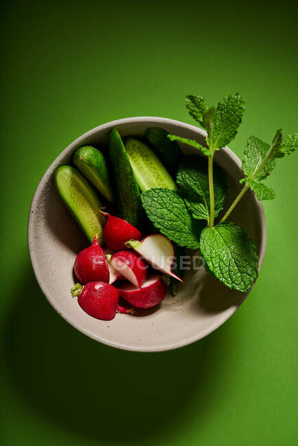 From above of cut fresh cucumbers with radish and leaves of mint placed in bowl served on green table — Stock Photo