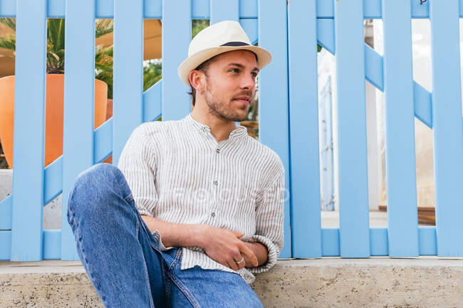 Schöner junger bärtiger Kerl in stylischem gestreiftem Hemd und Strohhut schaut weg, während er im Sommer am blauen Zaun chillt — Stockfoto