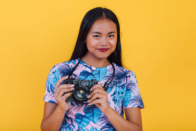 Glückliche asiatische Frau im T-Shirt mit tropischem Blatt-Print hält Fotokamera auf gelbem Hintergrund im Studio — Stockfoto