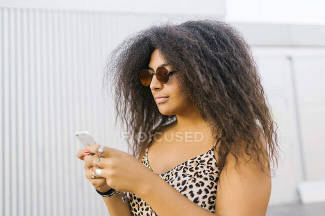 Mujer joven y afro con gafas de sol charlando con su teléfono inteligente - foto de stock