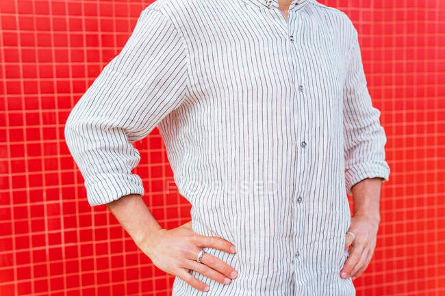 Crop unrecognizable bearded male in trendy casual striped shirt standing with hands on hip against red wall — Stock Photo