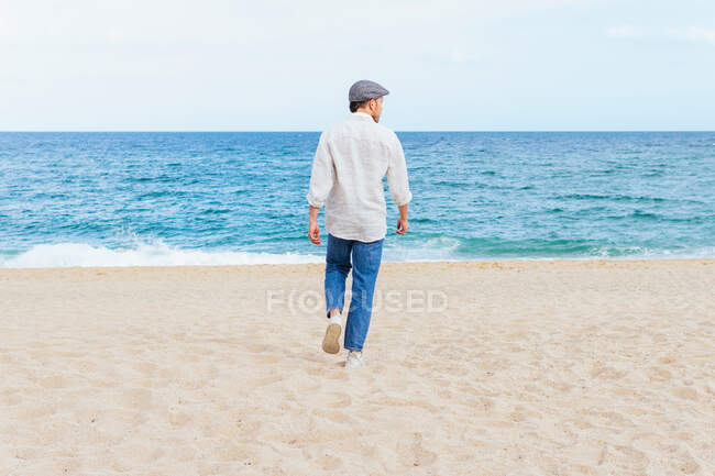 Vista trasera de cuerpo completo de un hombre irreconocible con ropa casual de moda y gorra caminando solo en la playa de arena hacia el mar ondeando mientras pasa las vacaciones de verano en la orilla del mar - foto de stock