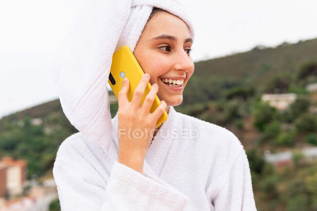 Молодая женщина в халате и полотенце улыбается и просматривает мобильный телефон во время отдыха на балконе после душа — стоковое фото