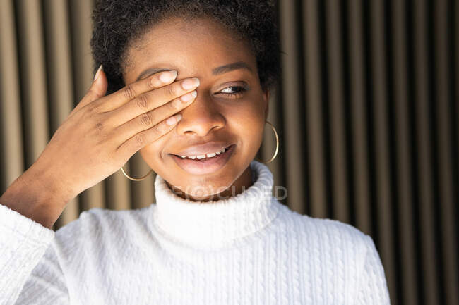 Happy African American female in trendy sweater covering eye and looking away with smile against striped wall on street — Stock Photo