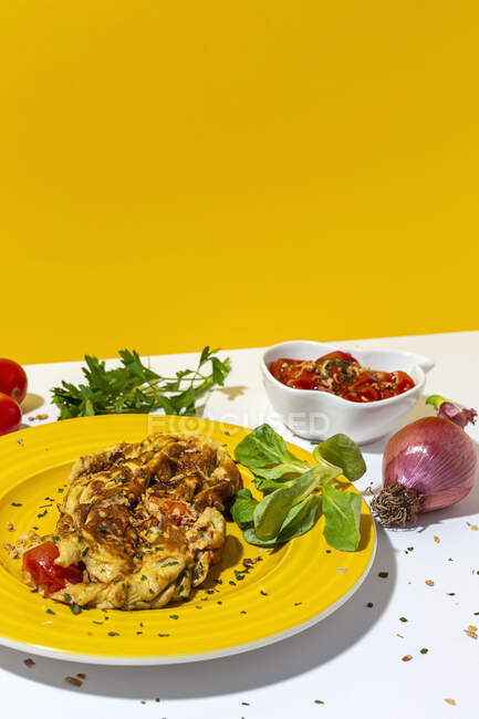 Deliciosa tortilla con perejil picado en plato contra tomates secados al sol y cebolla roja cruda sobre fondo blanco y amarillo - foto de stock