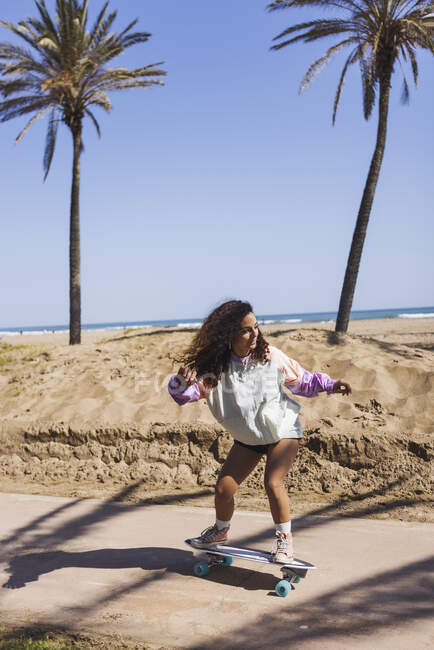 Corpo inteiro de fêmea ativa em roupas casuais andando de skate na estrada ao longo da praia de areia e palmas altas durante o treinamento — Fotografia de Stock