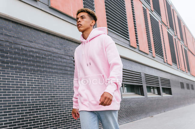 Angle bas de sérieux auto-assuré jeune homme ethnique avec des cheveux afro teints portant un sweat à capuche rose et un jean marchant près d'un bâtiment urbain moderne — Photo de stock