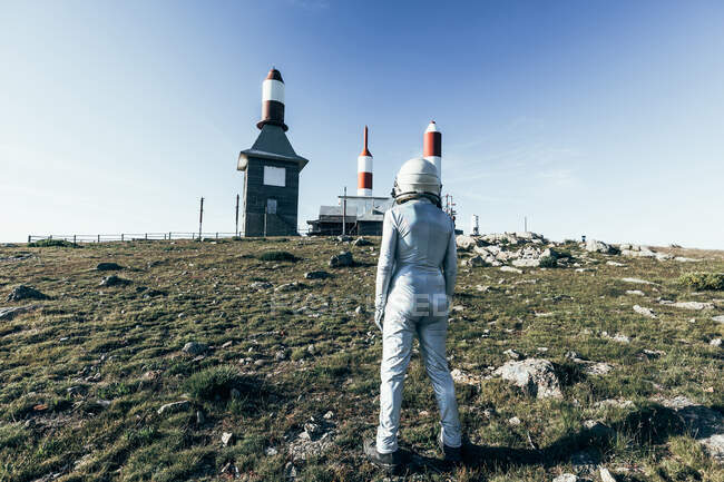 Rückansicht Ganzkörper-Mann im Raumanzug steht auf felsigem Boden gegen Metallzaun und gestreifte raketenförmige Antennen an sonnigen Tagen — Stockfoto