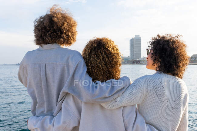 Visão traseira de amigos anônimos com cabelos encaracolados de pé perto no abraço contra a paisagem urbana e mar à luz do sol — Fotografia de Stock