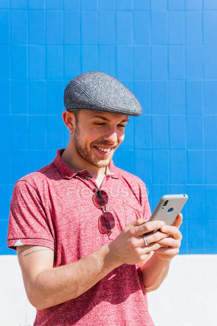 Contenido joven barbudo hipster chico en polo casual camisa y gorra de navegación teléfono móvil mientras que de pie contra la pared azul a la luz del sol - foto de stock