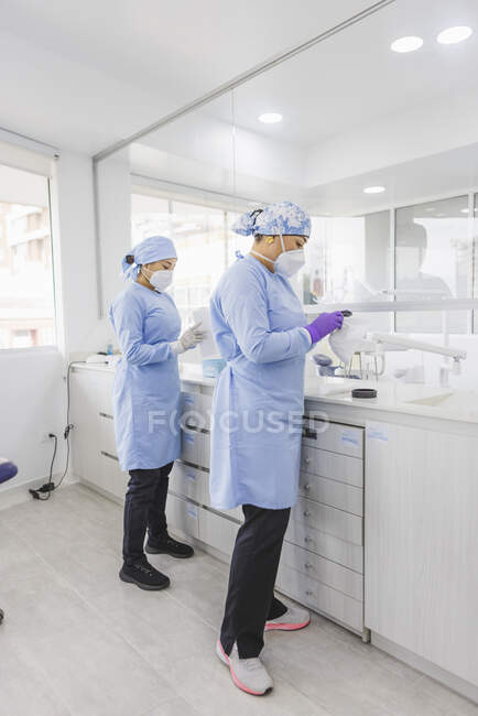 Vista lateral de los médicos femeninos en máscaras estériles de pie en la mesa con equipo profesional en la clínica - foto de stock