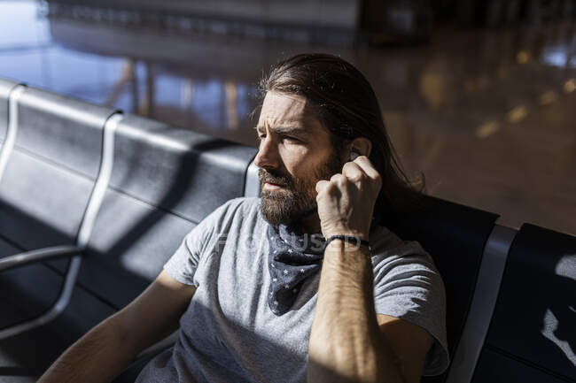 Der Mann mit dem Hut am Flughafen im Wartezimmer sitzt und wartet auf seinen Flug, er setzt sich kabellose Kopfhörer auf, um Musik zu hören — Stockfoto
