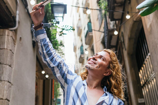 Beau mâle positif avec les cheveux longs prenant autoportrait sur smartphone tout en se tenant dans la rue en été — Photo de stock