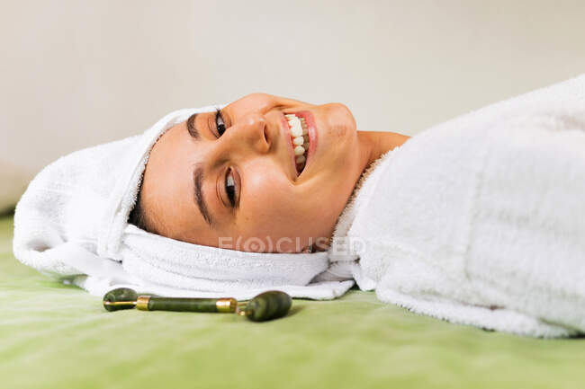 Vue de dessus jeune femme heureuse avec serviette sur la tête sourire et masser le visage avec rouleau de jade pendant la routine de soins de la peau à la maison — Photo de stock