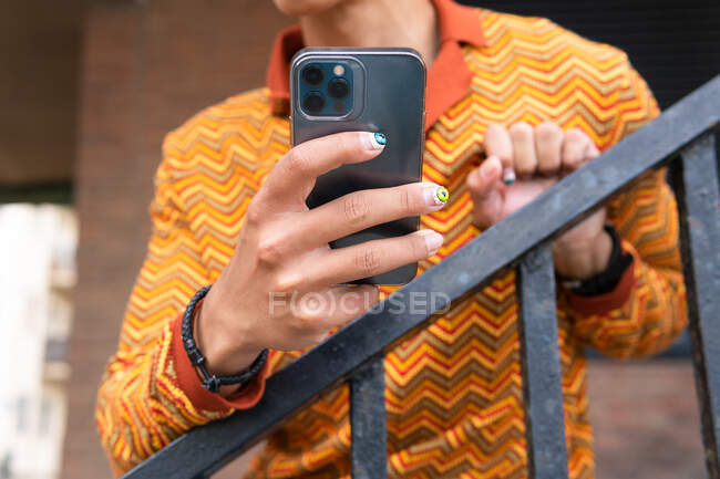 Crop unrecognizable stylish guy in fancy striped shirt with accessories and painted nails using mobile phone while standing near railing in city — Stock Photo