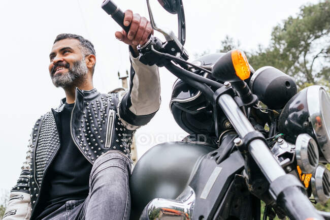 From below bearded ethnic male biker in black leather jacket sitting on modern motorbike on asphalt road amidst lush green trees growing in mountainous valley — Stock Photo