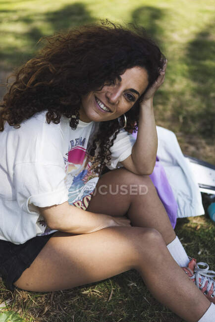 Positive female wearing white t shirt and shorts leaning on hand and looking at camera while sitting on grassy lawn — Stock Photo