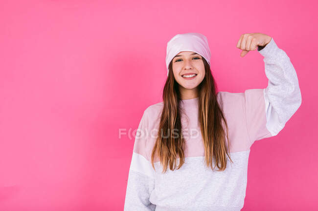 Contenida adolescente en traje casual y pañuelo para la cabeza para el concepto de cáncer demostrando brazos fuertes mientras mira a la cámara con sonrisa dentada - foto de stock