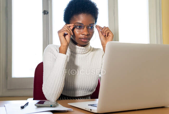 Mujer afroamericana joven concentrada en ropa casual y anteojos mirando la pantalla del portátil y leyendo información profesional mientras trabaja en la mesa en la oficina - foto de stock