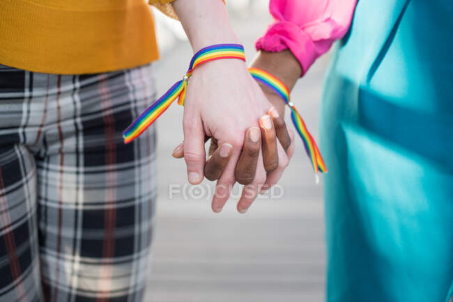 Cultivez un couple multiethnique méconnaissable de lesbiennes avec des bracelets arc-en-ciel LGBT tenant la main en ville — Photo de stock