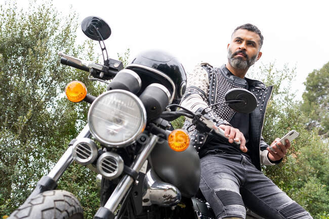 Low angle of concentrated adult brutal ethnic male biker with beard in leather jacket with rivets sitting on modern motorbike and looking away while messaging on smartphone — Stock Photo