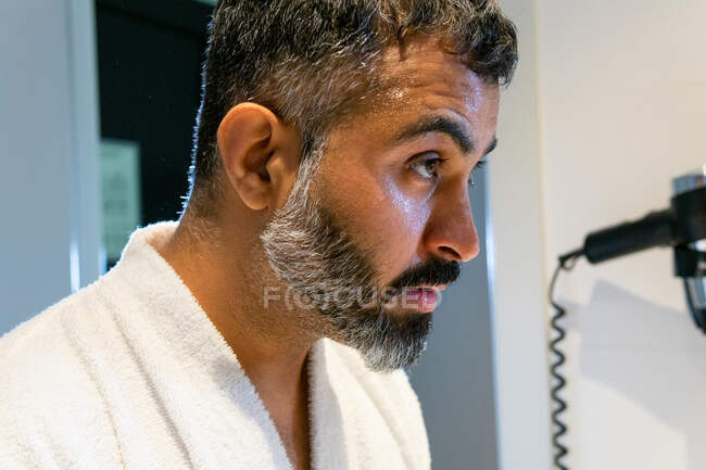 Vista laterale del raccolto maschio di mezza età in accappatoio bianco faccia di pulizia con acqua durante la routine mattutina in bagno — Foto stock