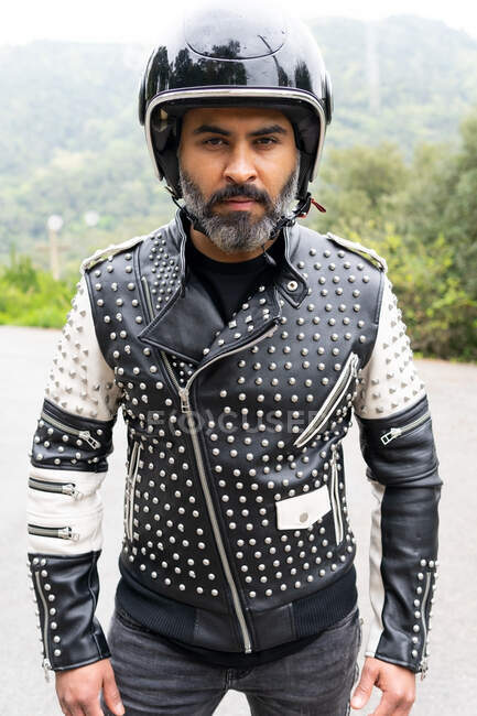 Homme hispanique d'âge moyen barbu veste en cuir à la mode avec rivets et casque noir regardant la caméra tout en se tenant debout sur la route près d'arbres verts luxuriants dans la nature — Photo de stock