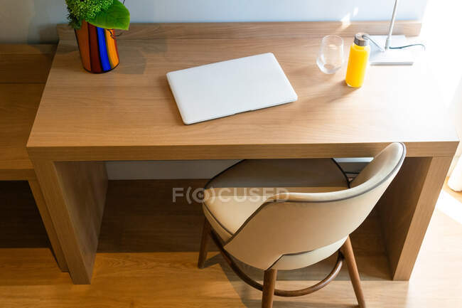 Удобное кресло рядом со столом со стеклянной бутылкой свежего сока в номере и ноутбуком рядом с цветком — стоковое фото