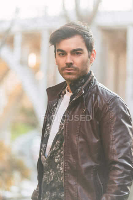 Homem latino confiante vestindo jaqueta de couro na moda em pé no parque de outono e olhando para a câmera — Fotografia de Stock