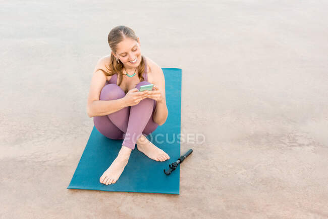 Задоволена жінка в активному одязі, сидячи на йога мат, переглядаючи на мобільному телефоні на березі моря — стокове фото