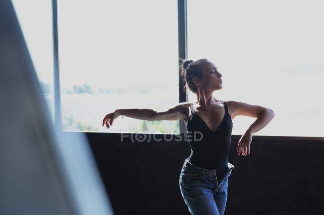 Jovem fêmea em jeans com cabelo bun dançando enquanto olha para o chão com sombras à luz do sol — Fotografia de Stock