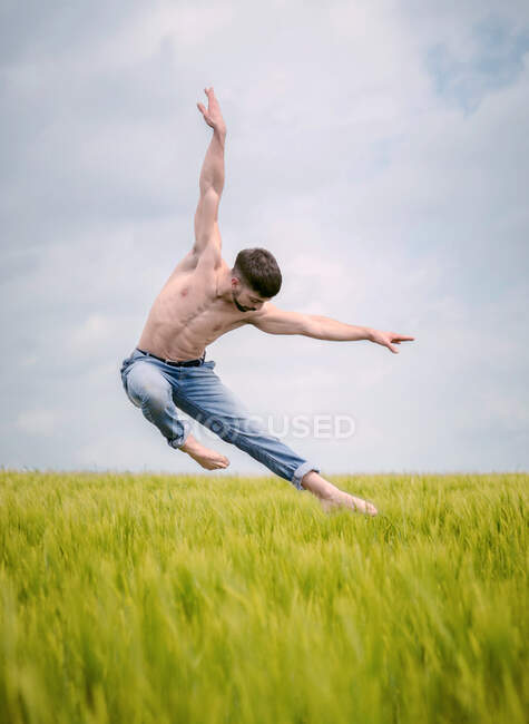 Shirtless man in denim performing sensual ballet jump with arms spread above tall grass in gloomy field — Stock Photo
