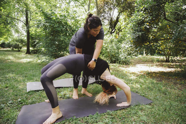 Тренер дорослих допомагає невпізнаваній жінці стояти в позі Урдхви Дханурасани під час тренування йоги в літньому парку. — стокове фото