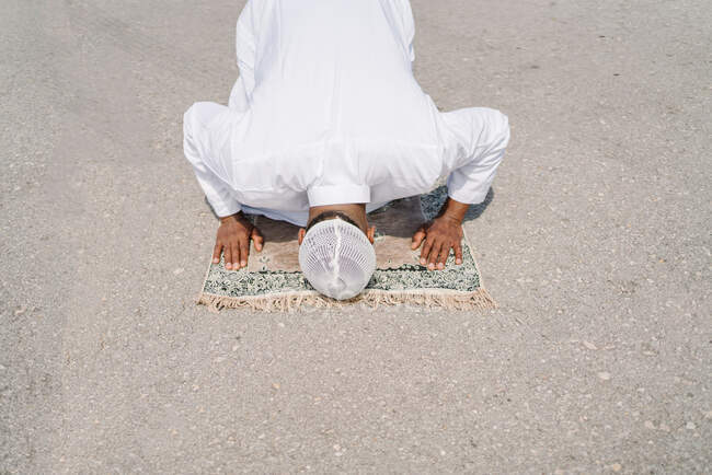 Unrecognizable Muslim male kneeling on rug and touching ground with forehead while praying on sandy beach on sunny day — Stock Photo