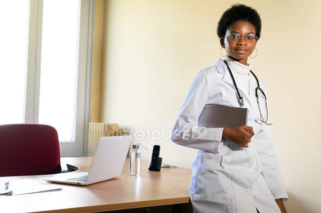 Позитивная молодая черная женщина-врач в медицинском халате и очках со стетоскопом, смотрящая в камеру во время работы с планшетом в современном офисе клиники — стоковое фото