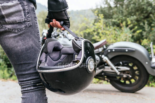 Back view of crop anonymous male biker in jeans and leather gloves holding helmet in hand while standing on asphalt road near parked modern motorcycle — Stock Photo