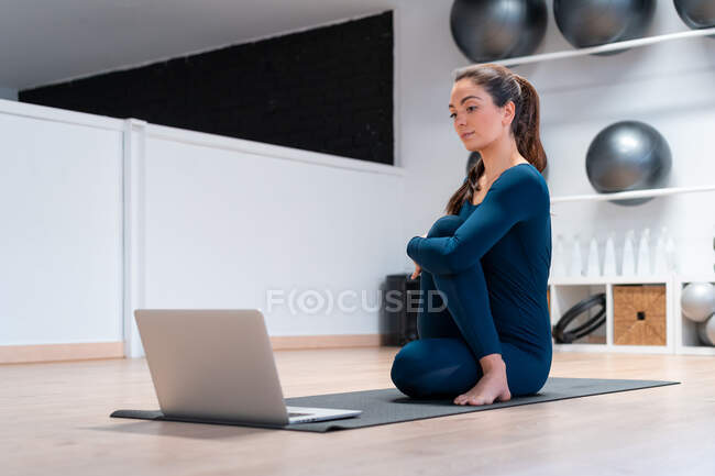 Cuerpo completo de instructora de yoga joven y flexible haciendo la pose de mitad señor de los peces frente a la pantalla del portátil durante la clase en línea en el gimnasio - foto de stock